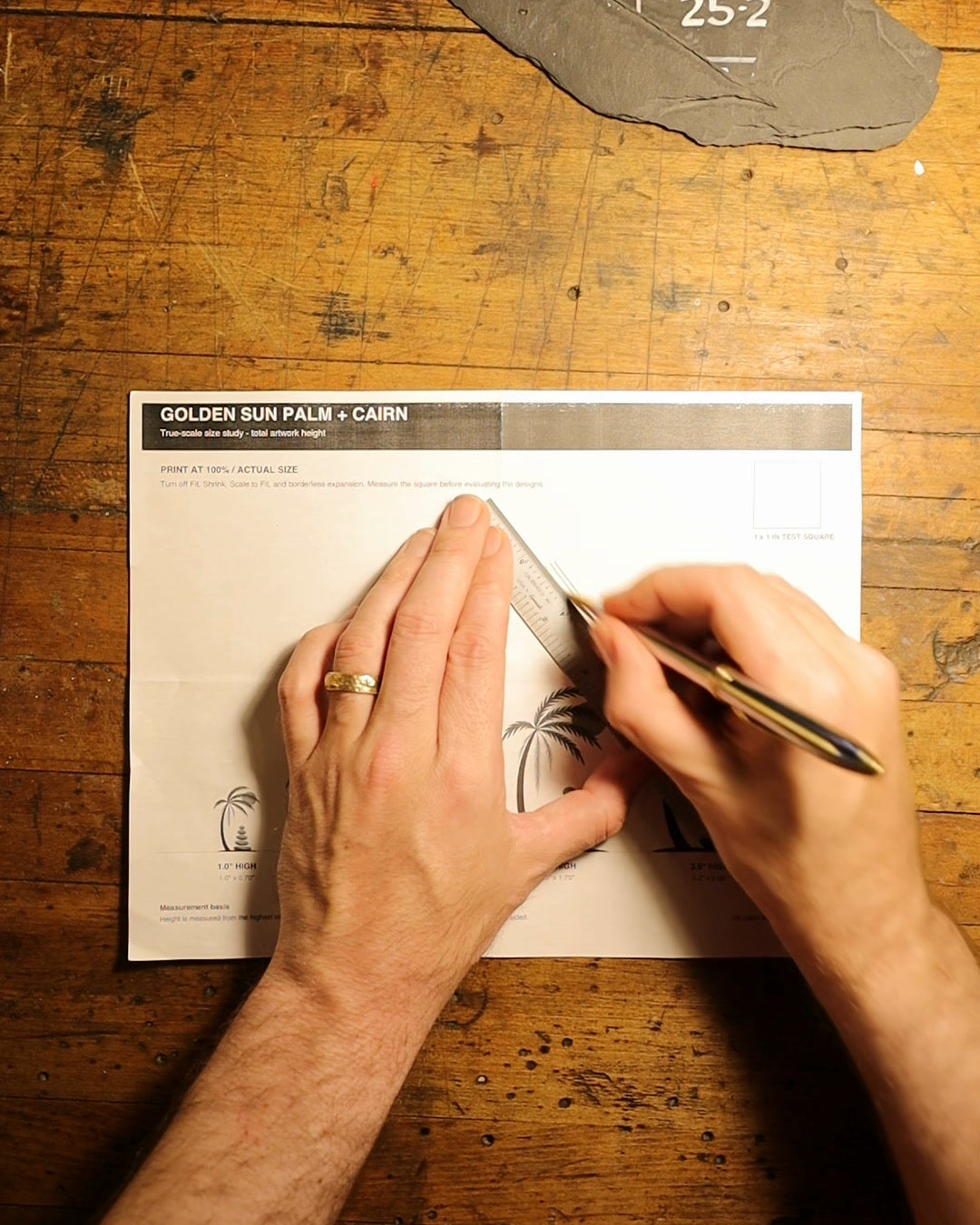Hands using a ruler and pen on an Inspired Outdoors palm-and-cairn design study at the workbench.